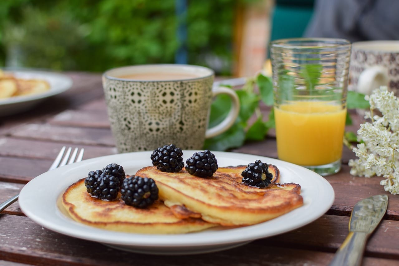 Pancakes topped with blackberries placed on an outdoor dining table in front of a coffee mug and a glass or orange juice