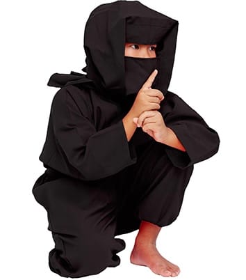 A child in a black ninja costume squats and holds their index finger to their lips in a shushing gesture