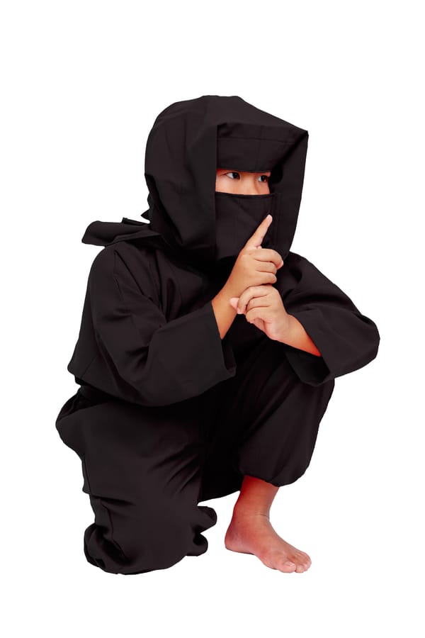 A child in a black ninja costume squats and holds their index finger to their lips in a shushing gesture